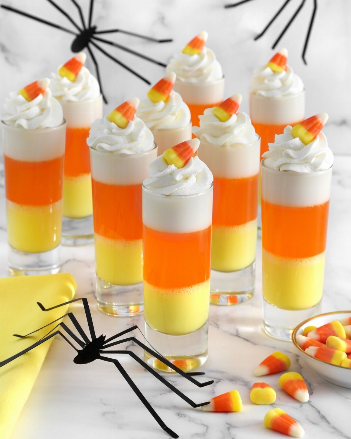 Layered yellow and orange jello shots topped with whipped cream and candy corn on marble.