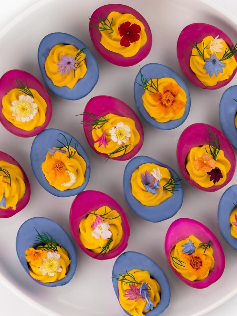 Dyed deviled eggs with pink and blue shells topped with yellow yolk and edible flowers.