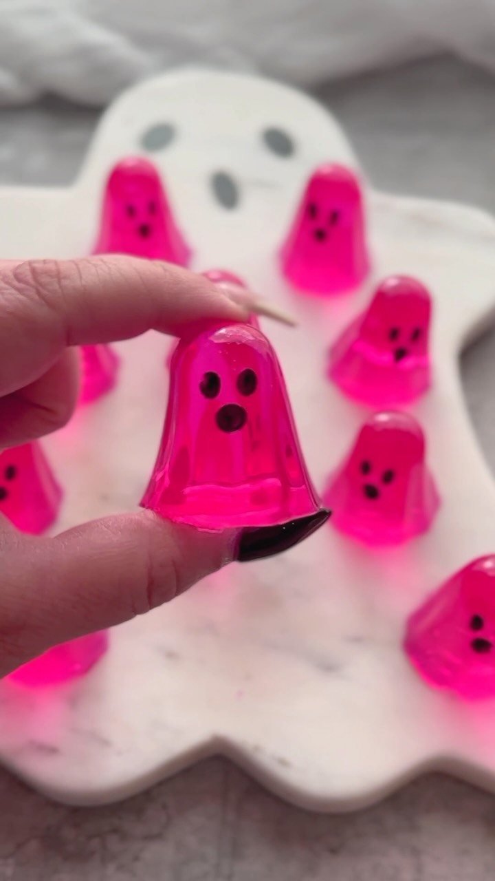 A hand holds up a bright pink ghost-shaped jello shot over a marble platter.