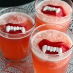 Three glasses of red frothy punch topped with floating vampire teeth on a lace cloth.