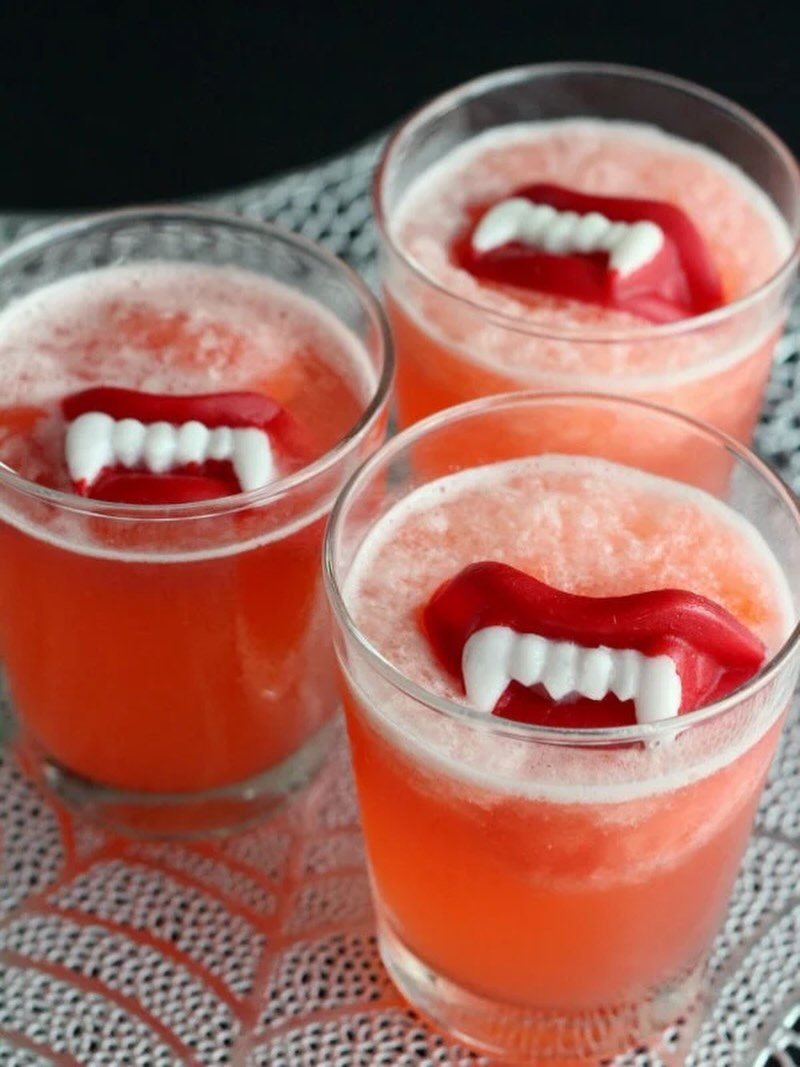 Three glasses of red frothy punch topped with floating vampire teeth on a lace cloth.