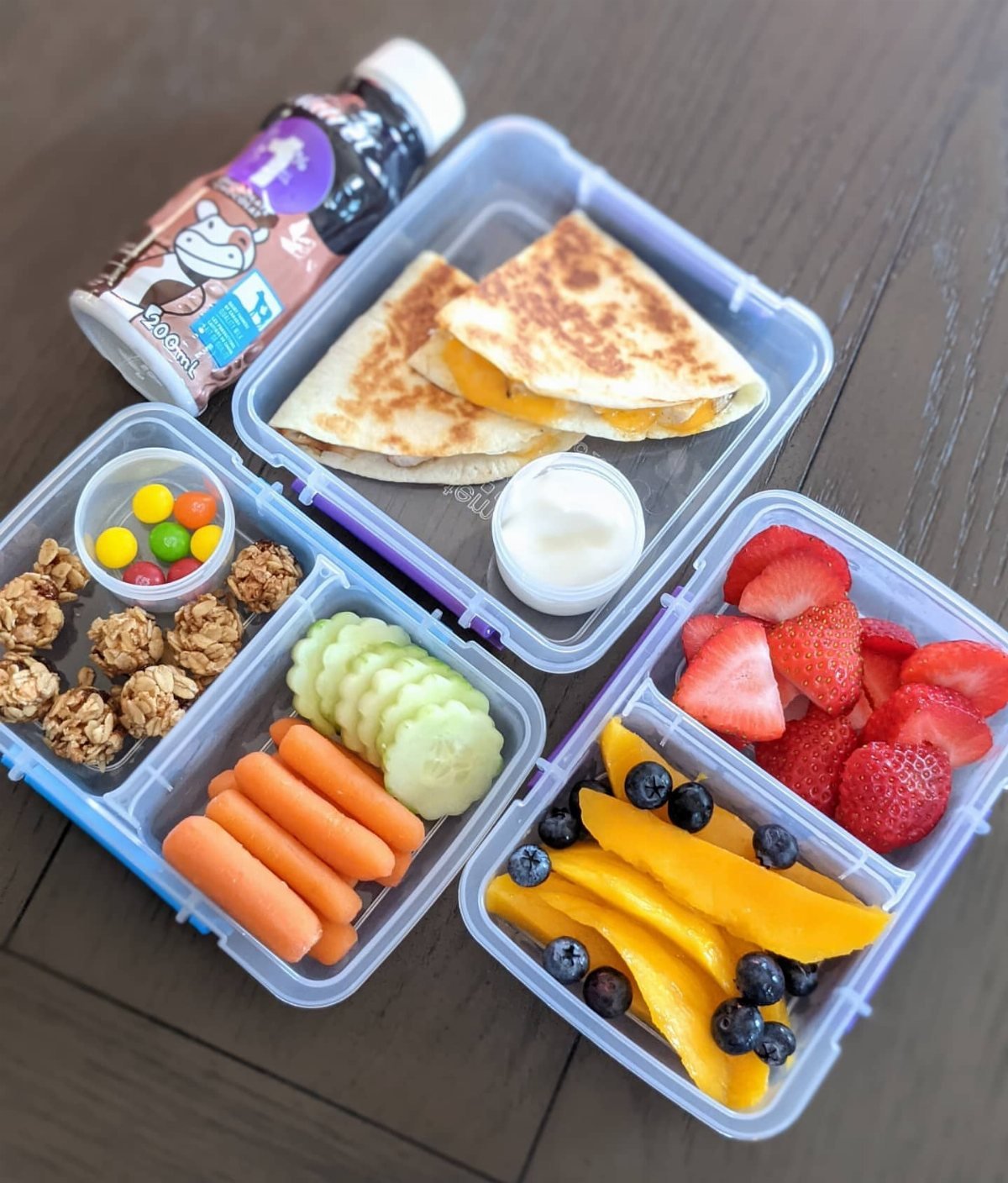Three bento lunch boxes containing quesadillas, vegetables, granola, and fresh fruit on a wooden table.