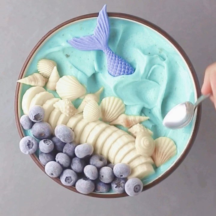 Blue smoothie bowl topped with blueberries, banana slices, seashells, and a purple mermaid tail.