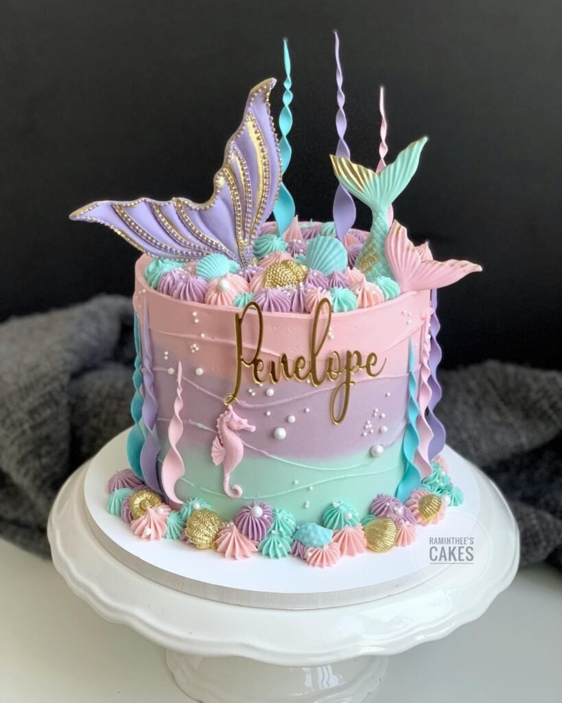 A pastel mermaid themed cake with seashell frosting and gold name lettering on a white stand.